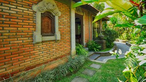 Um edifício de tijolos tradicional com uma porta de madeira decorativa, rodeado por vegetação exuberante e um caminho de pedra.