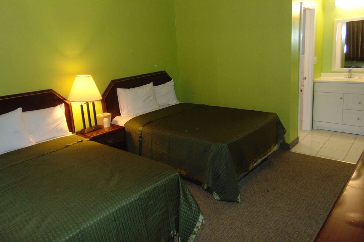 A hotel room with two beds, a nightstand with a lamp, a bathroom with a sink and mirror, and green walls.
