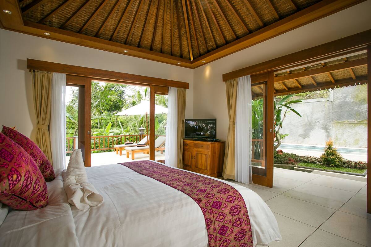 A bedroom with a large bed, a television, and a view of a pool and garden.