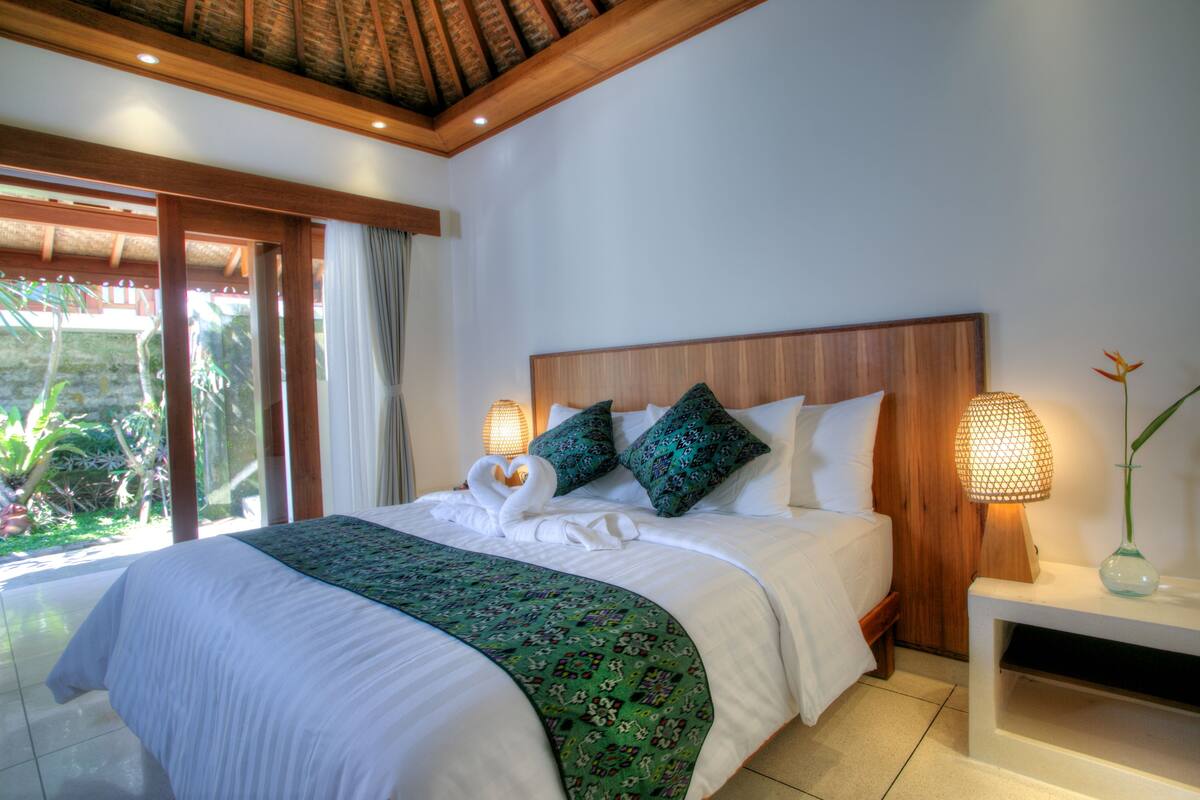 A neatly made bed with a green patterned bedspread, a wooden headboard, and two decorative pillows. There is a bedside lamp on a white nightstand, and a window with curtains allowing natural light.