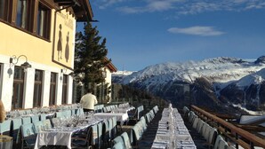 A long dining table set for a meal with mountain views in the background. - Hotel Salastrains (St. Moritz)