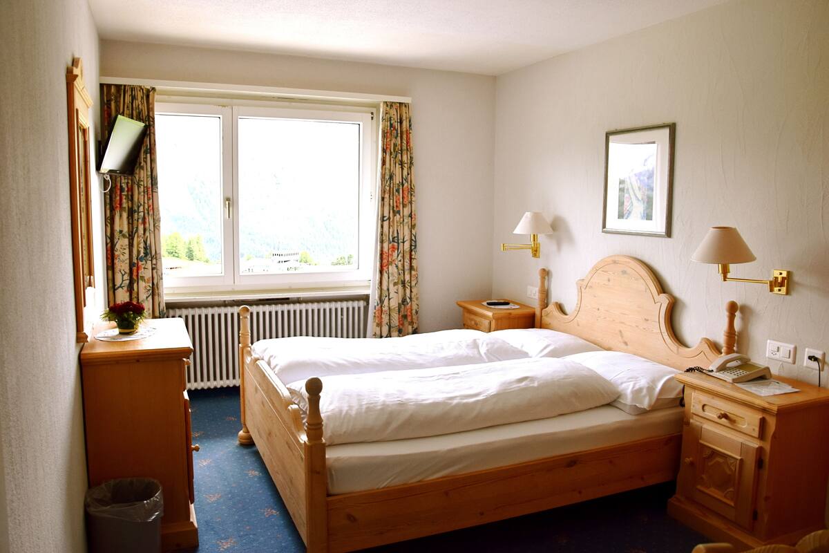 A bedroom with a wooden bed, a bedside table with a lamp, a radiator, a window with curtains, and a small trash bin.