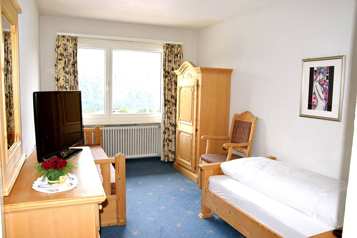 A room with a bed, a wooden chair, a desk with a television and flowers, a radiator, and a window with curtains.