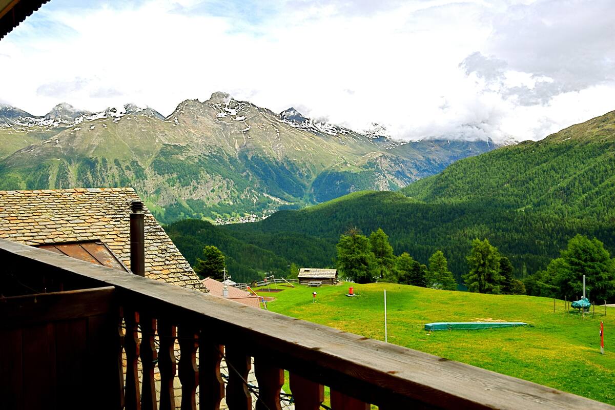A mountain view with a wooden balcony, a stone-roofed building, and a green field.