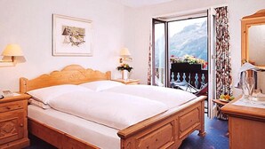 A wooden bed with a white mattress, two bedside tables with lamps, a framed picture on the wall, and a view of mountains through an open door. - Hotel Salastrains (St. Moritz)
