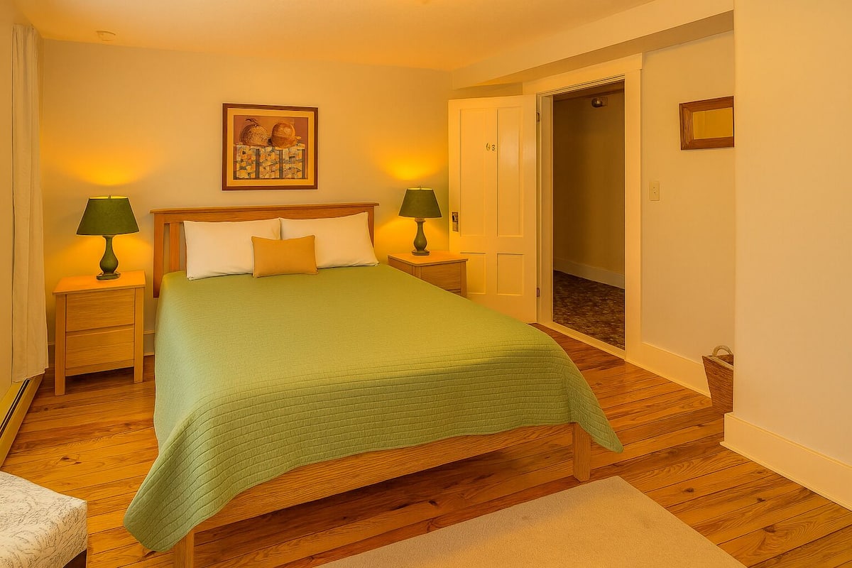 A neatly arranged bedroom with a large bed, two bedside tables with lamps, a framed picture on the wall, and a door leading to another room.