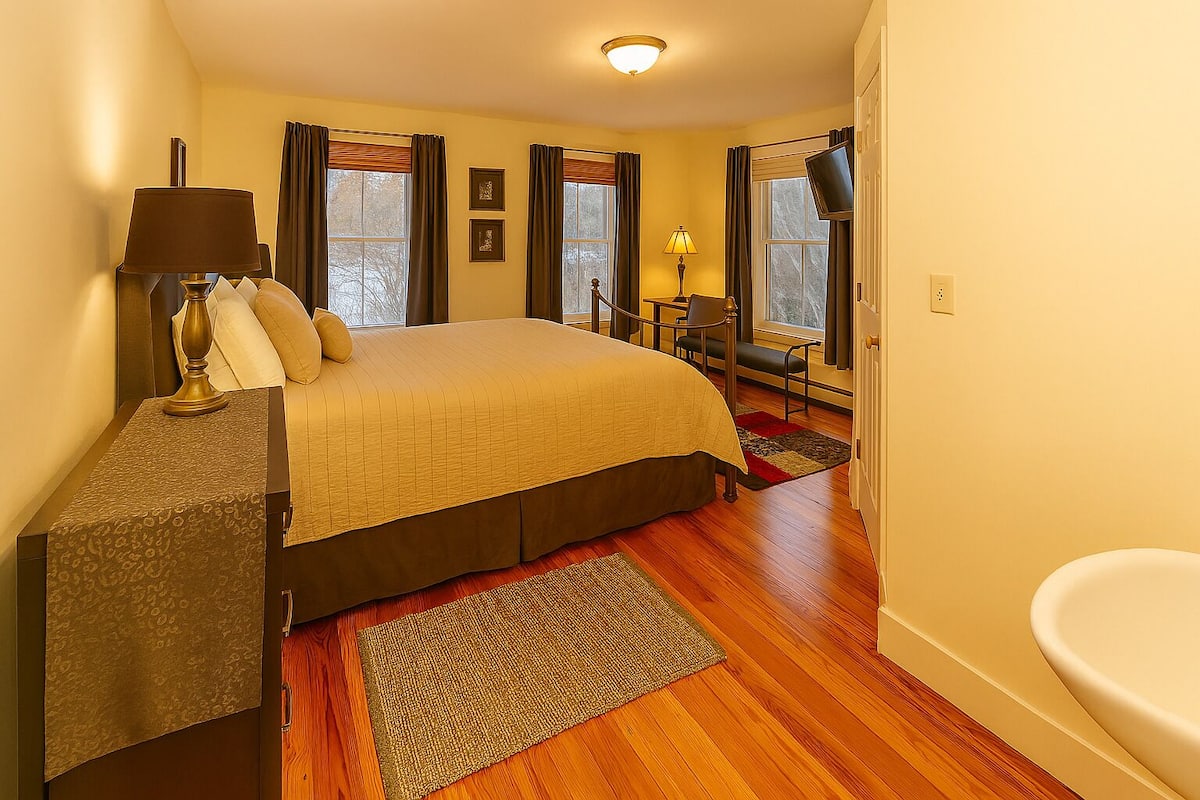 A bedroom with a bed, bedside table, lamp, and a view of the outside through windows.