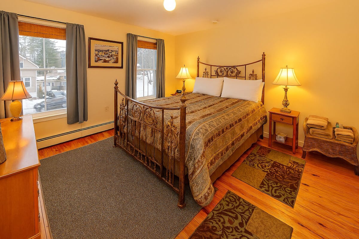 A bedroom with a wooden bed, two bedside lamps, a wicker chair, and a view of the outside.