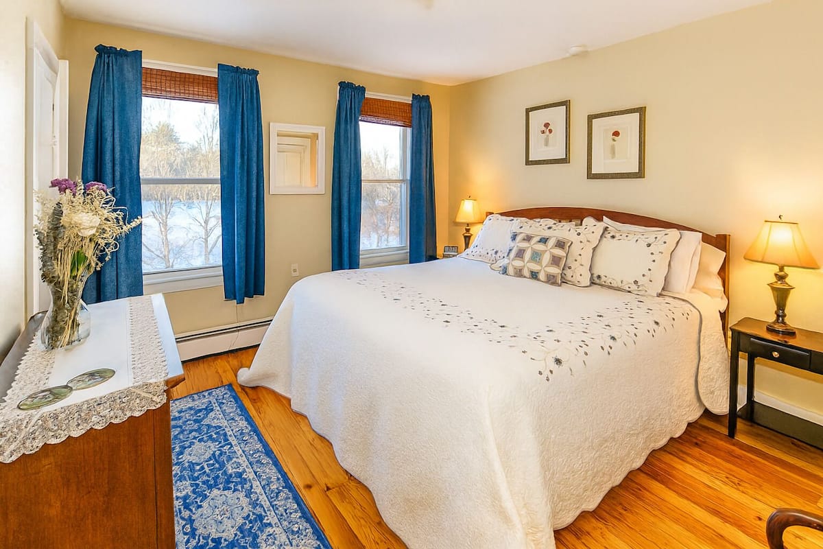 A bedroom with a large bed, a bedside table with a lamp, a vase with flowers, and a window with blue curtains.