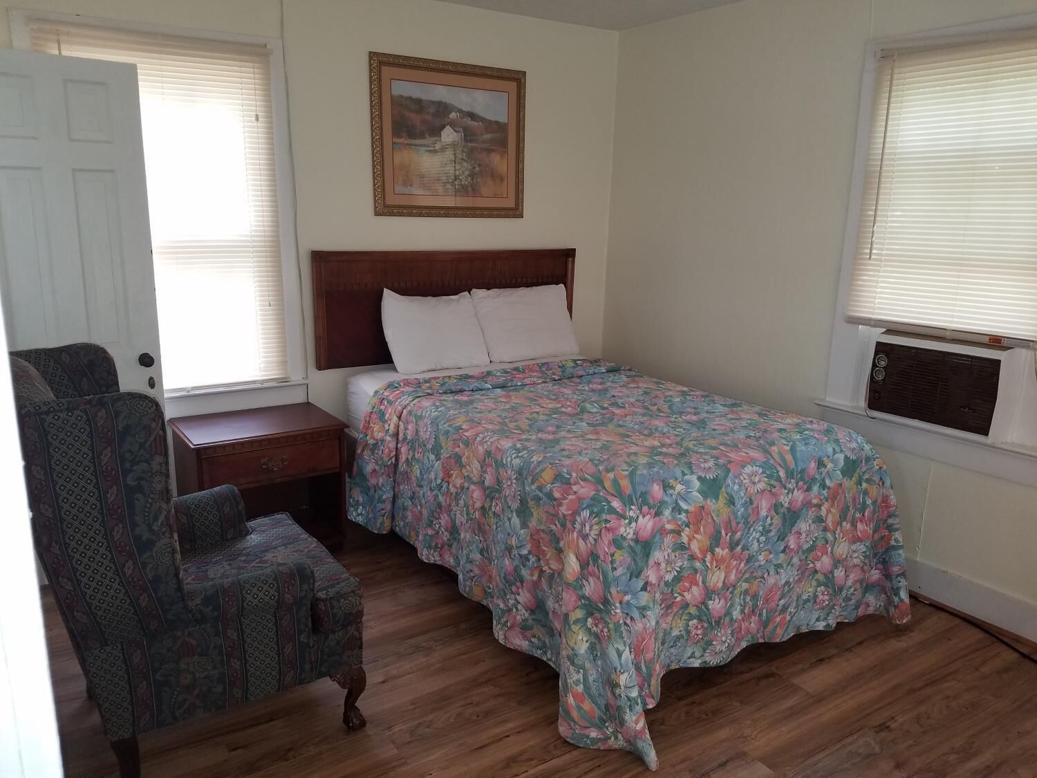 A bedroom with a bed, a chair, a nightstand, a painting on the wall, and an air conditioning unit.