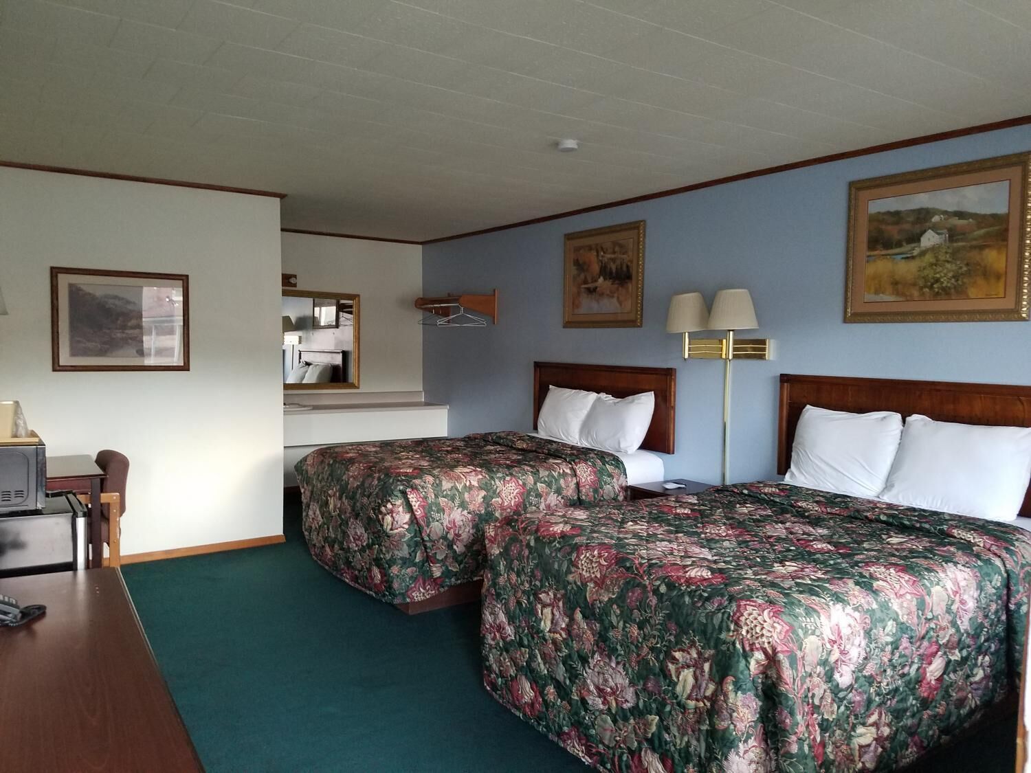A hotel room with two beds, a desk, a chair, a lamp, and artwork on the walls.