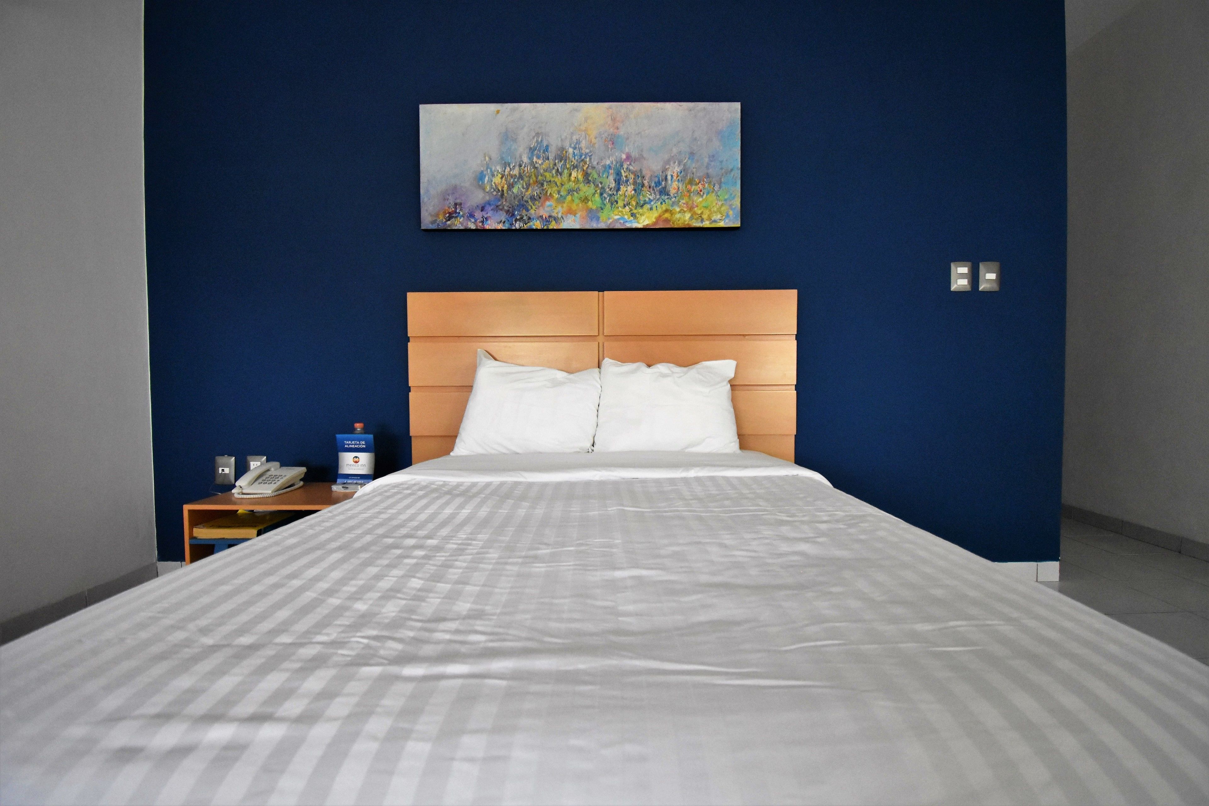 Quarto de hotel com uma cama grande, cabeceira de madeira, um quadro na parede, mesa de cabeceira com telefone e uma parede azul de destaque.