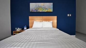 Quarto de hotel com uma cama grande, cabeceira de madeira, um quadro na parede, mesa de cabeceira com telefone e uma parede azul de destaque.