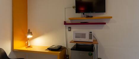 Una habitación pequeña con escritorio, silla, televisor, microondas y un bote de basura.
