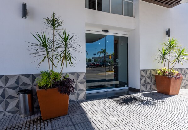Entrada de hotel moderna com paredes de padrão geométrico, plantas em vasos e vista desobstruída da rua.