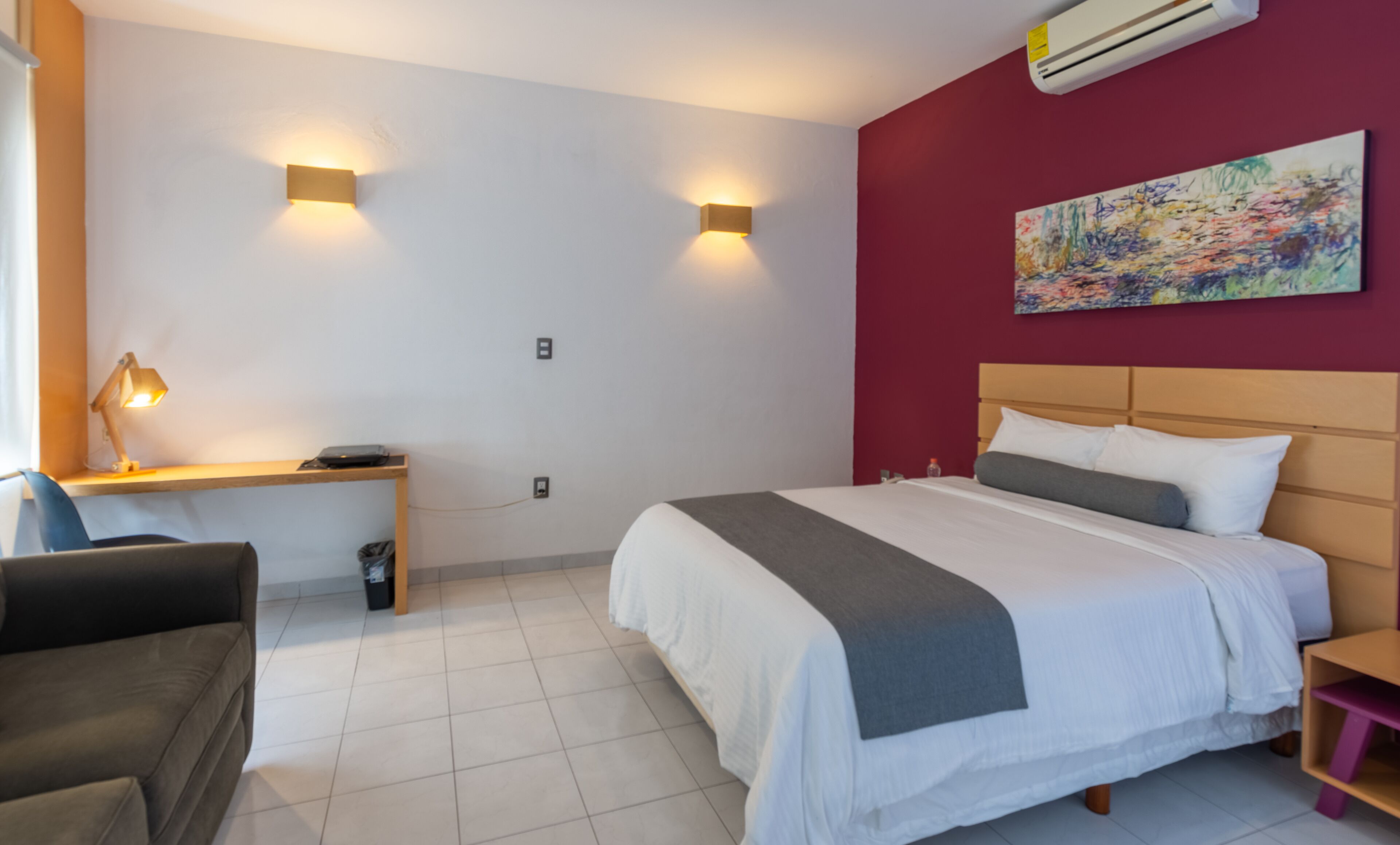 A hotel room with a bed, desk, chair, and a painting on the wall.