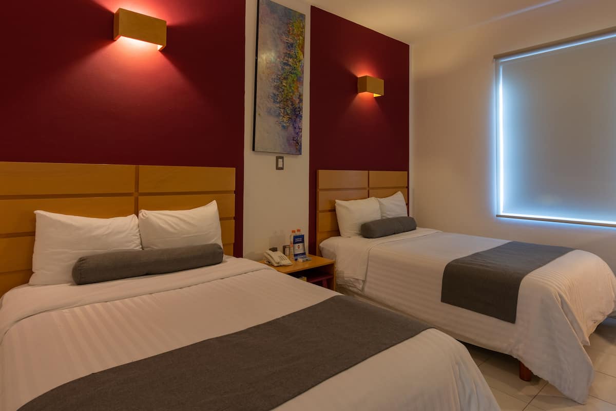 A hotel room with two beds, each with white bedding and gray throw pillows. There are wall-mounted light fixtures and a painting above the beds.