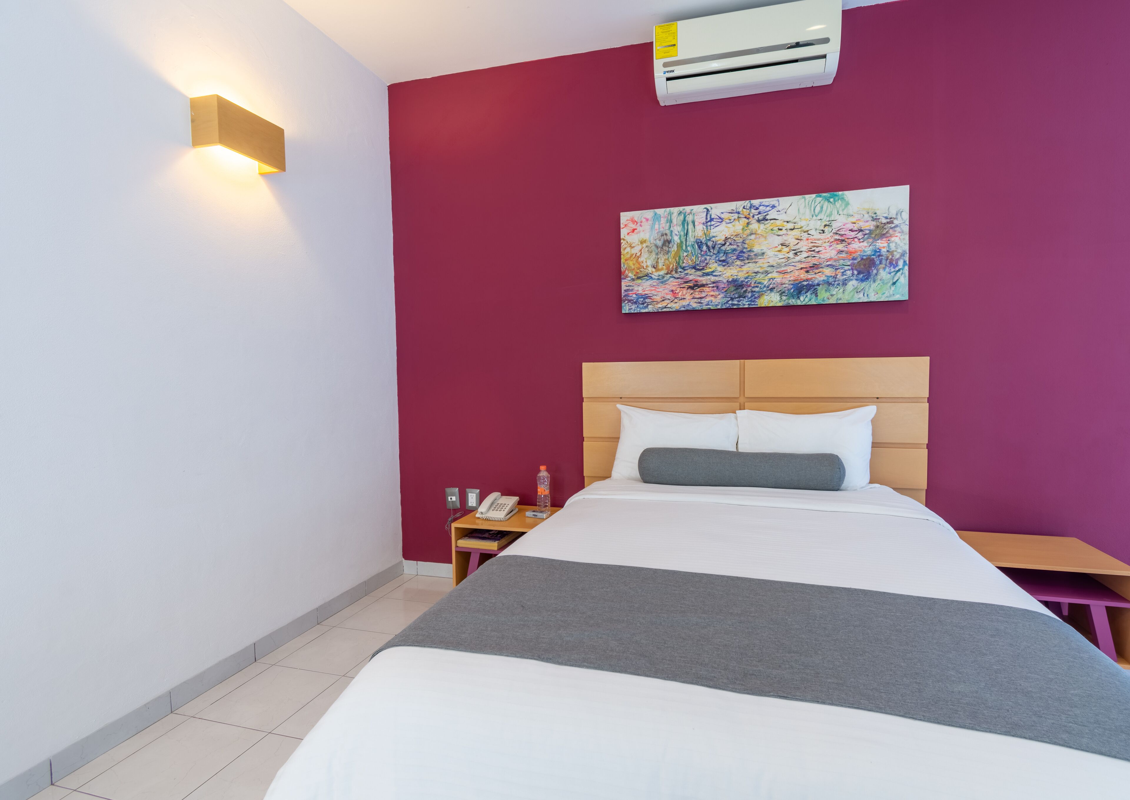 Ein Hotelzimmer mit einem Bett, einem Nachttisch, einem Telefon, einer Wasserflasche, einer Klimaanlage und einem Bild an der Wand.