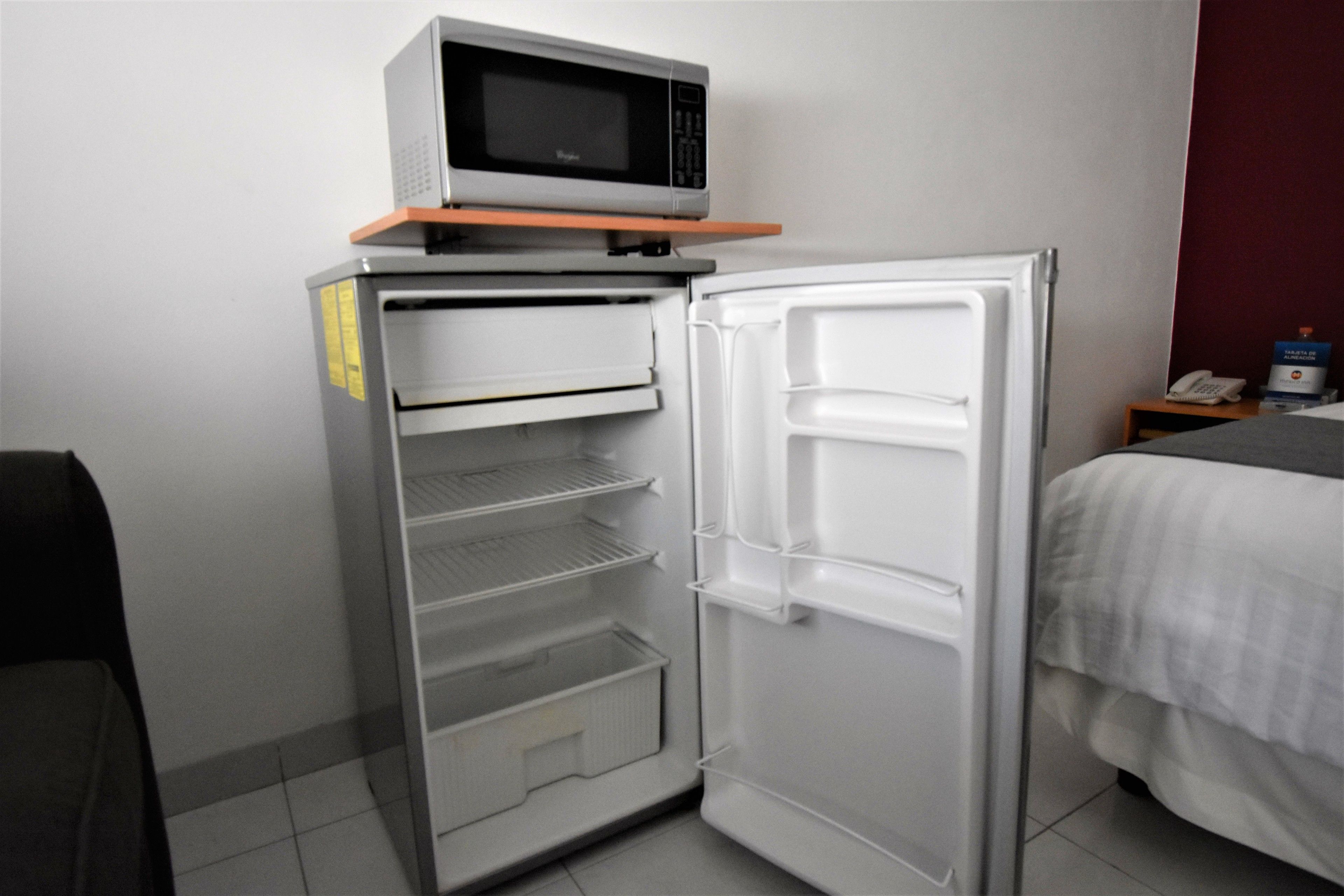 Una habitación de hotel compacta con refrigerador, microondas y una mesita de noche pequeña con teléfono y una caja.