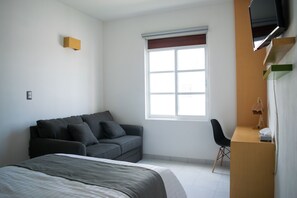 A bedroom with a bed, a grey sofa, a desk with a chair, a TV mounted on the wall, and a window with blinds.