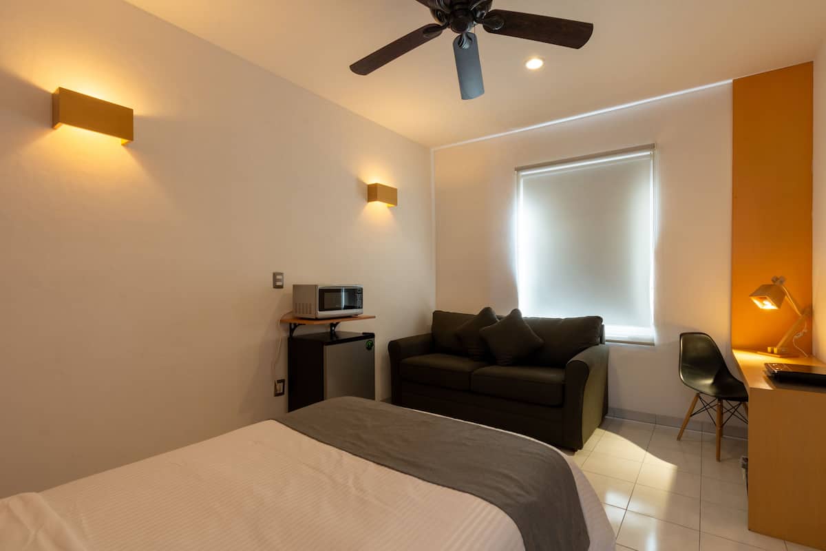 A modern hotel room with a bed, a sofa, a desk with a lamp, a ceiling fan, and a window with blinds.