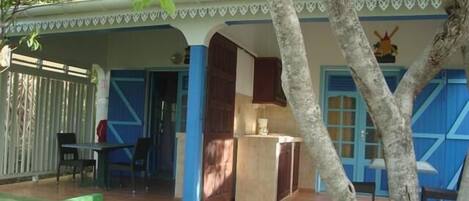Un espacio exterior techado con una mesa y sillas de madera, una pequeña área de cocina y una puerta azul.