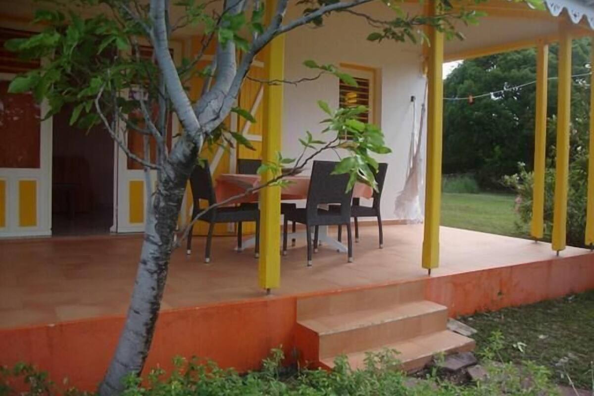 Un espacio al aire libre cubierto con una mesa y sillas, rodeado de pilares amarillos y un árbol en primer plano.