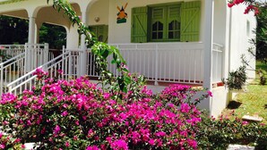 Um edifício branco com persianas verdes, um guarda-corpo branco e flores cor-de-rosa em primeiro plano.