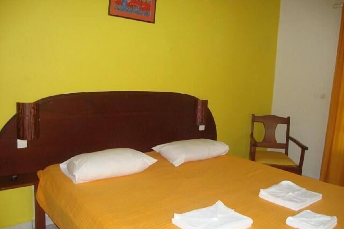 Una cama con cabecera de madera, dos almohadas blancas y una colcha naranja. Hay dos toallas blancas dobladas sobre la cama.