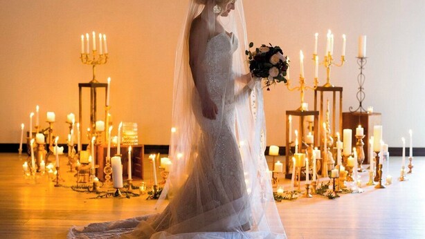 A bride in a white wedding dress holding a bouquet, standing in a spacious hall with wooden flooring and multiple lit candles arranged in rows. - Suites at The Red Rock Vacation rentals (Hot Springs)