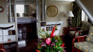 Una habitación acogedora con piso de madera, una cama con ropa de cama blanca, una estantería, un sofá estampado y una planta decorativa.