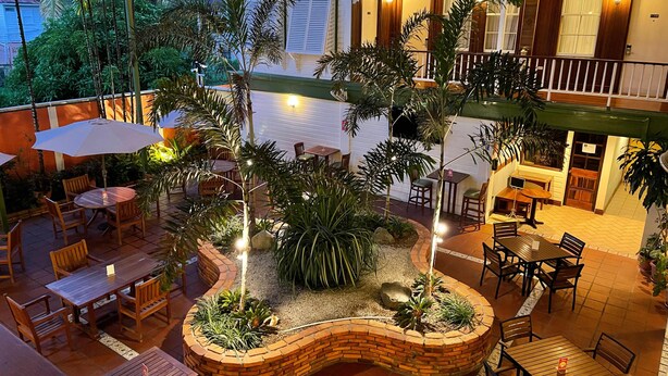 Un área de patio al aire libre con mesas y sillas de madera, rodeada de plantas y un macetero de ladrillo.