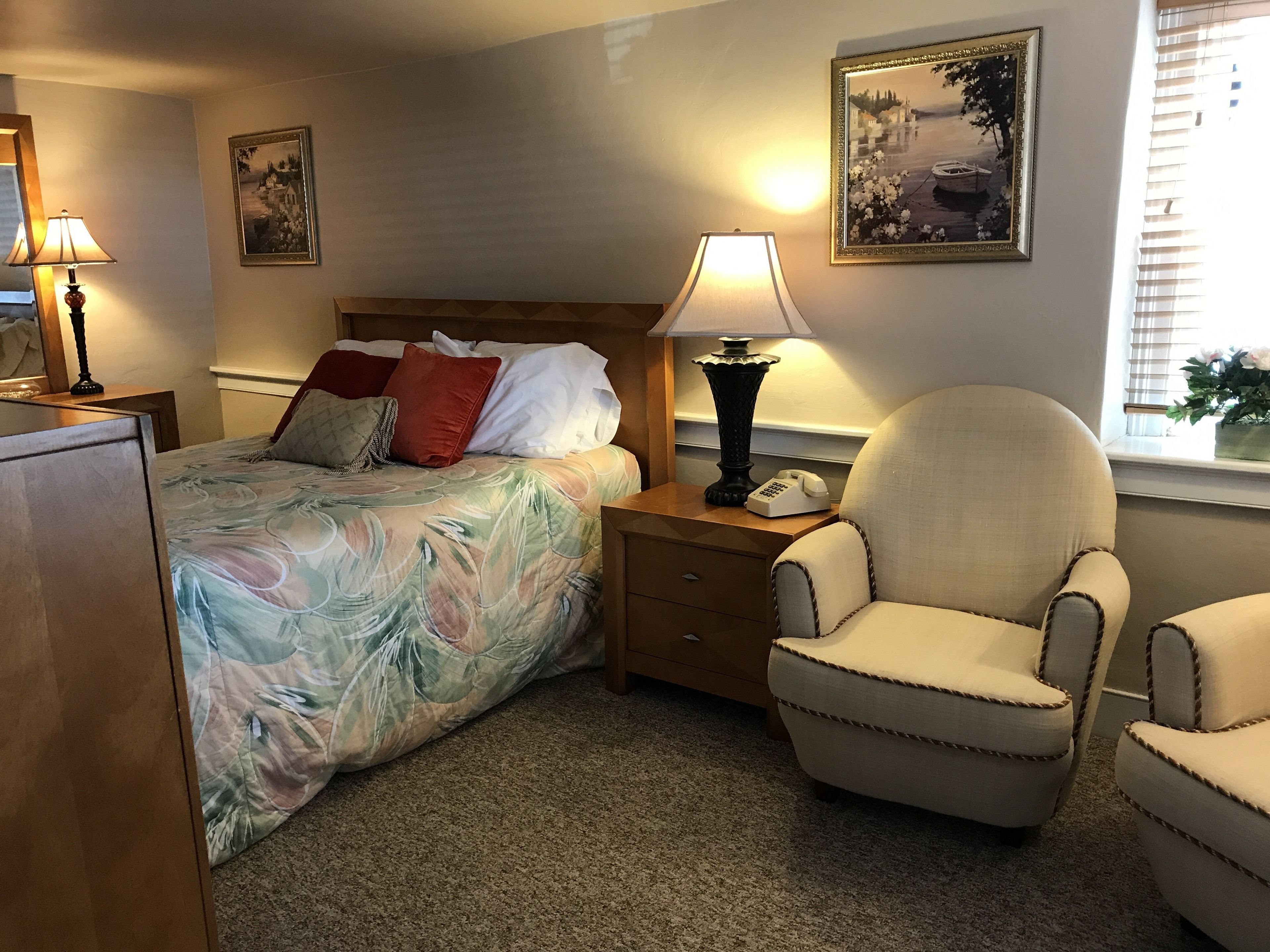 Quarto de hotel com cama, duas mesas de cabeceira, uma cadeira, abajur e um quadro na parede.