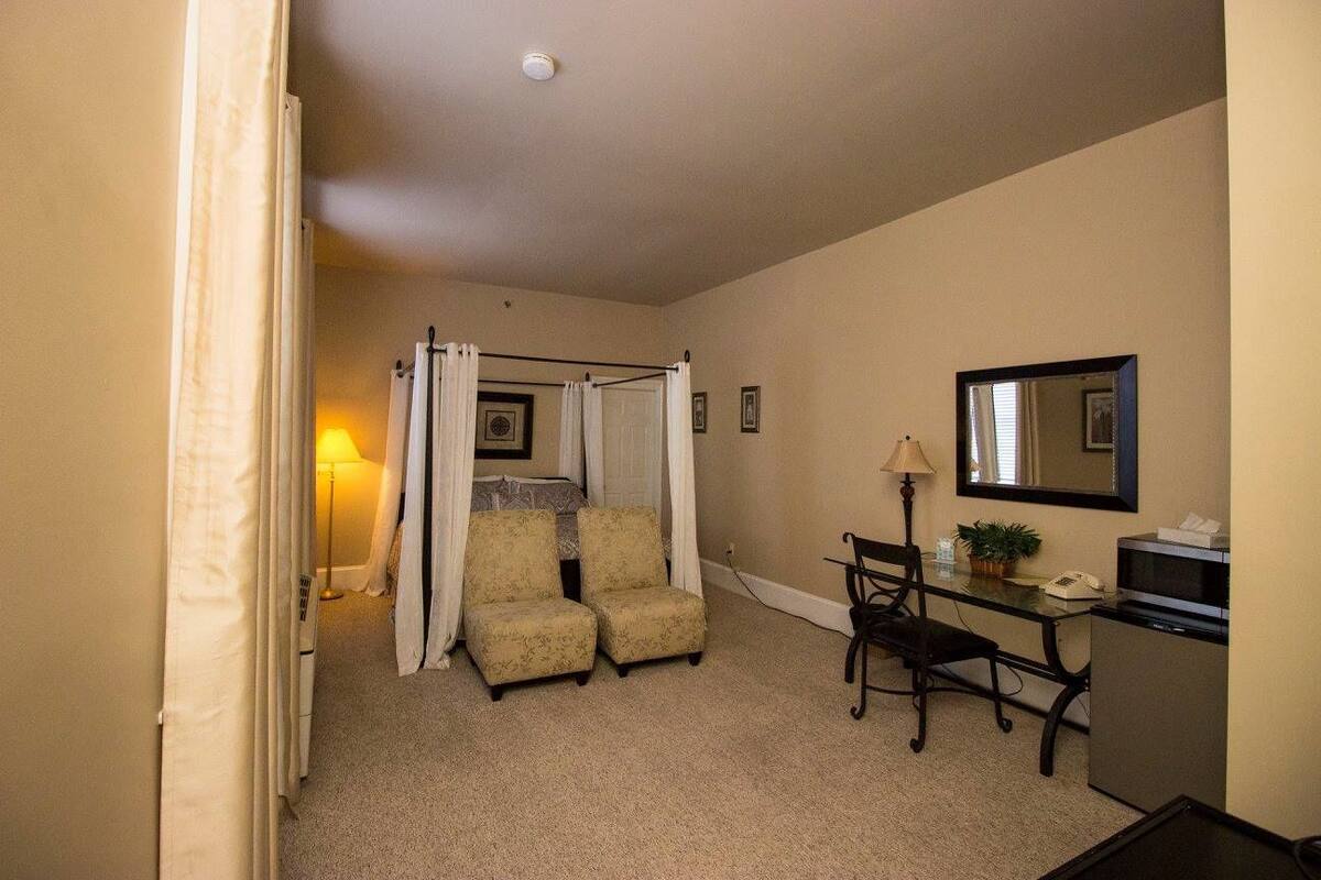 A hotel room with a bed, a desk with a chair, a small kitchenette, and a bathroom with a shower curtain.