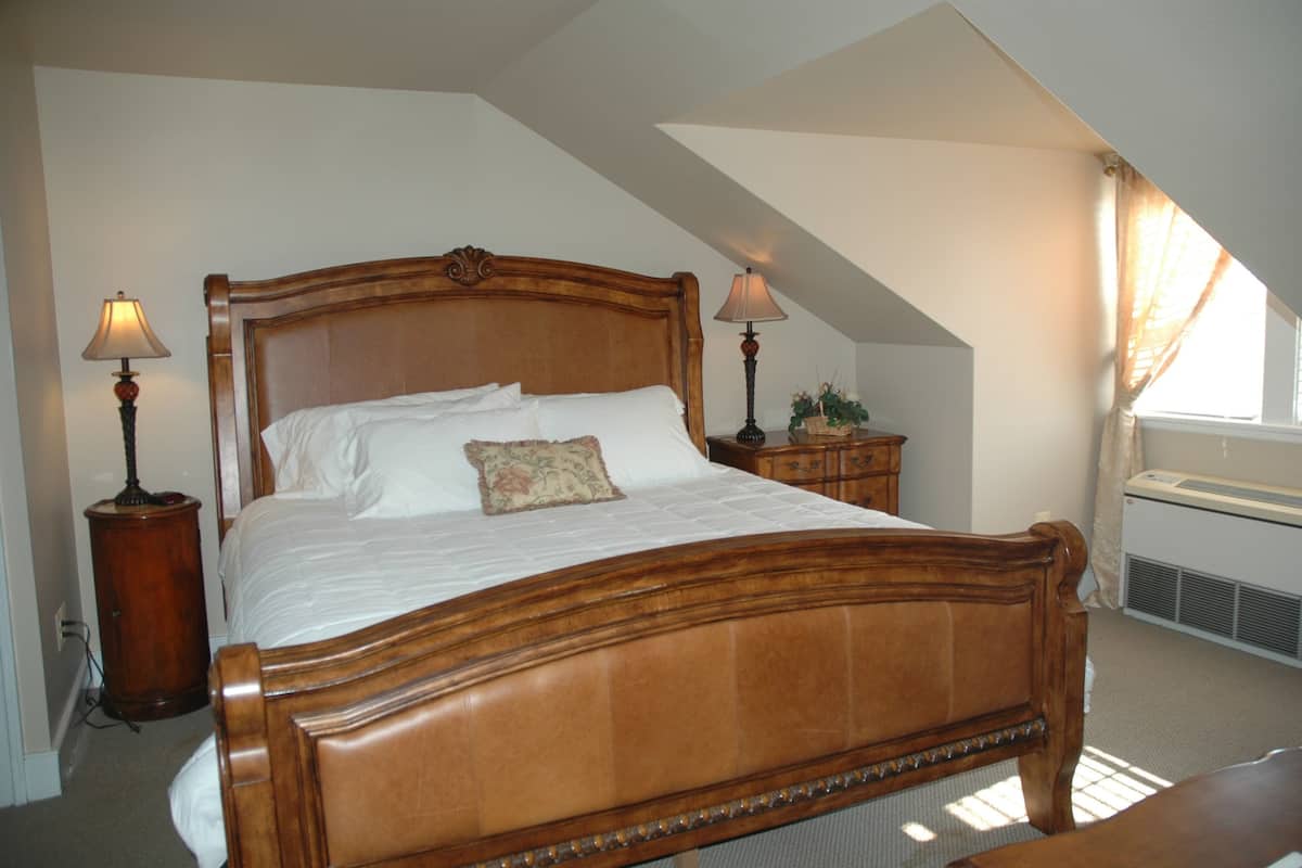 A wooden four-poster bed with white linens, two bedside tables with lamps, a potted plant, and an air conditioning unit.