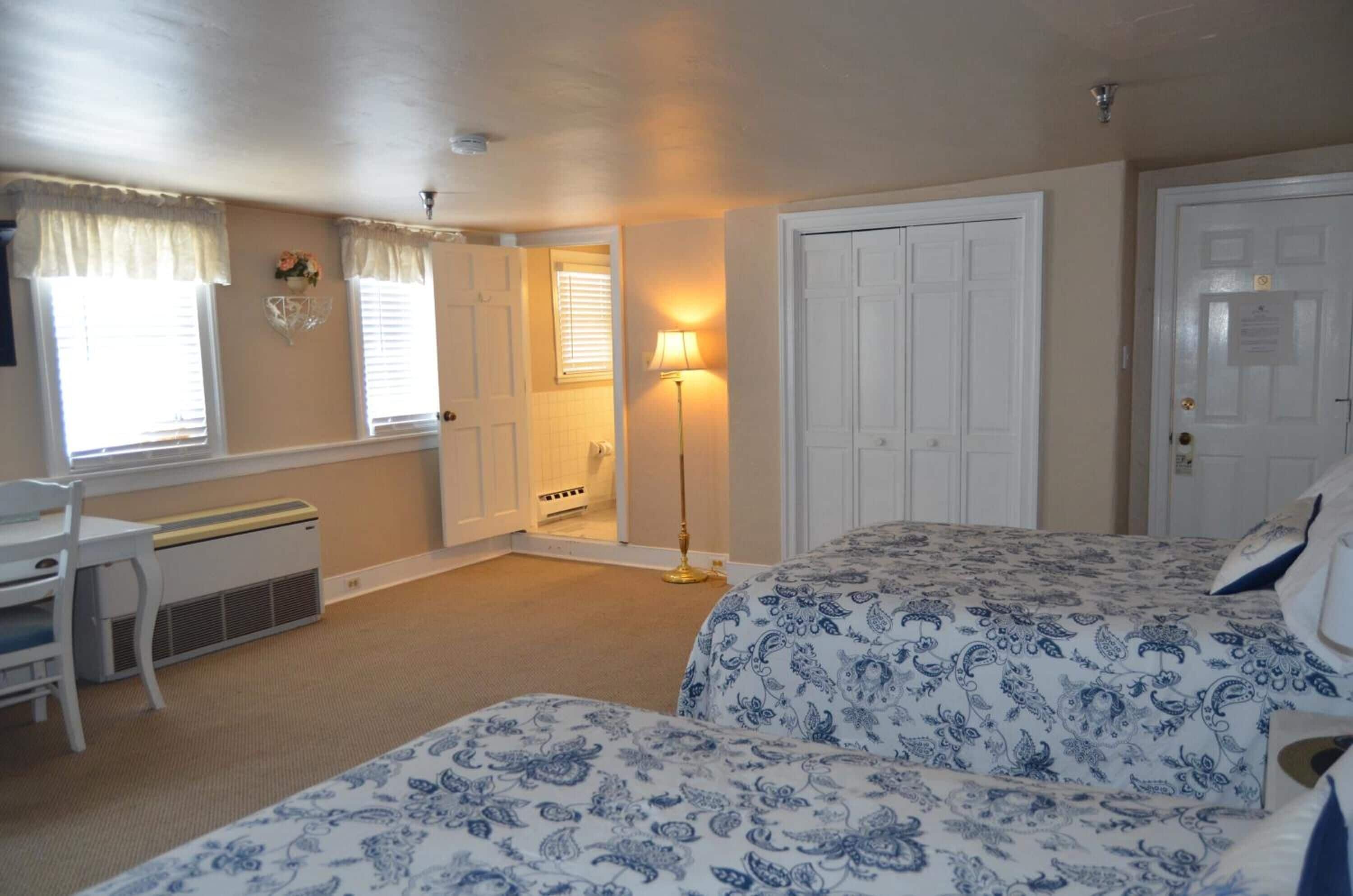 A bedroom with two beds, a desk, and a chair. There is a door leading to another room and a lamp in the corner.
