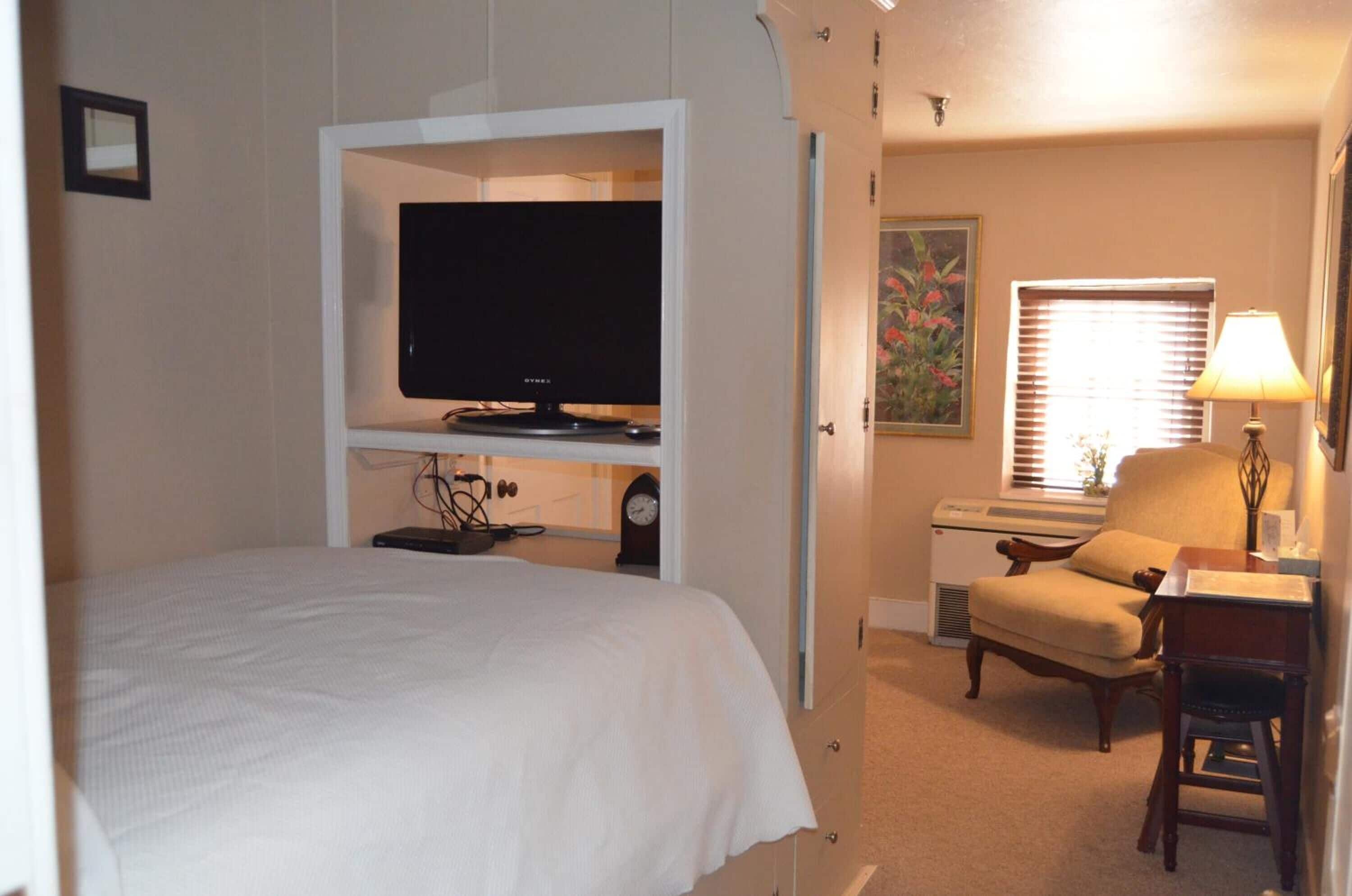 A hotel room with a bed, a television, a chair, a desk, and a window with blinds.