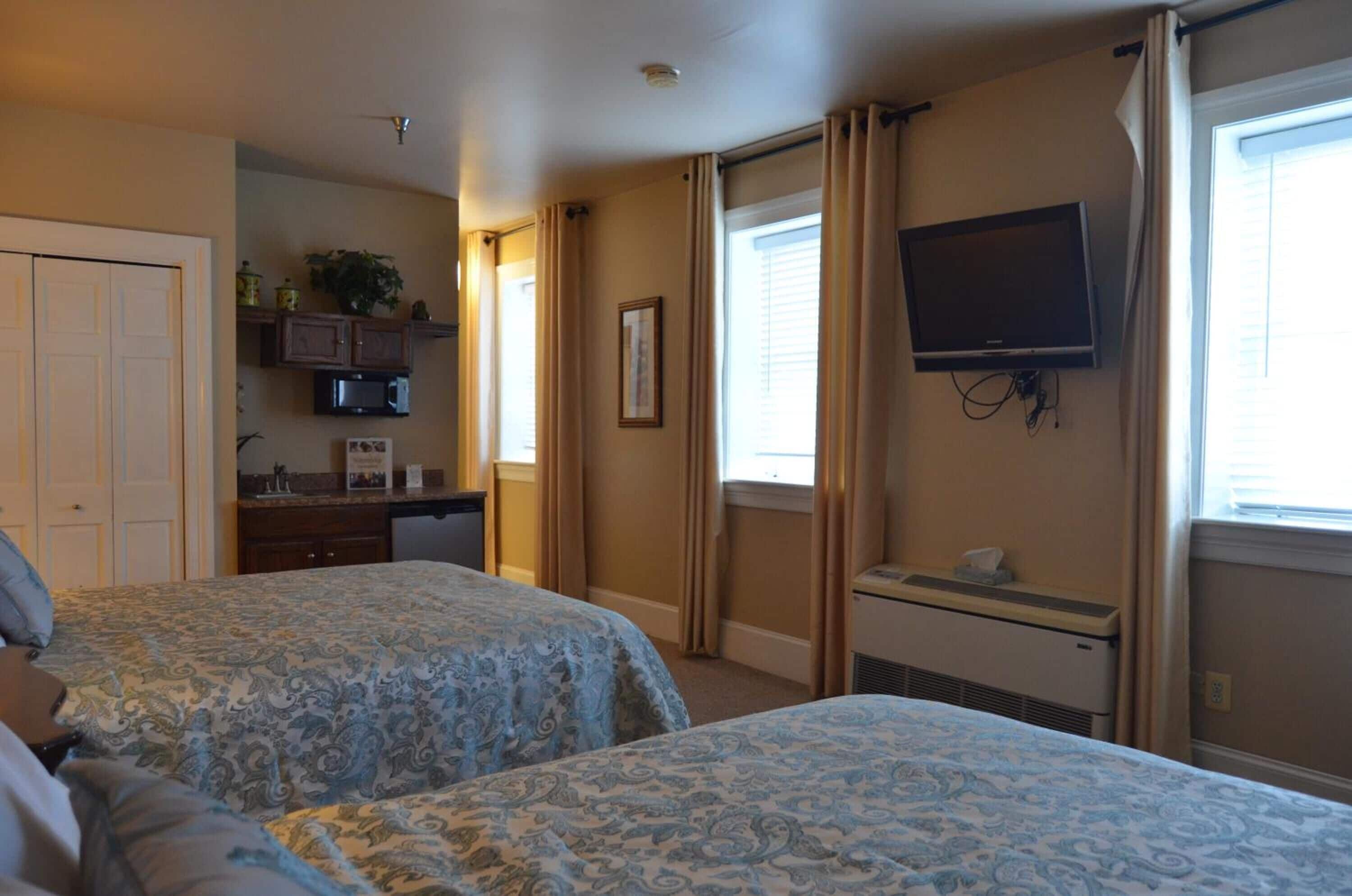 A hotel room with two beds, a TV mounted on the wall, a kitchenette, and a window with curtains.