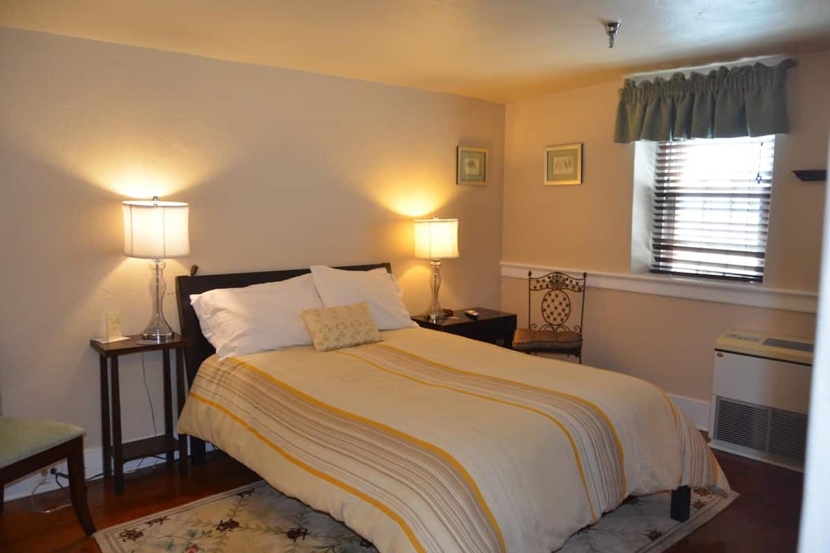 A neatly arranged bedroom with a bed, two bedside tables with lamps, a window with blinds, and a radiator.