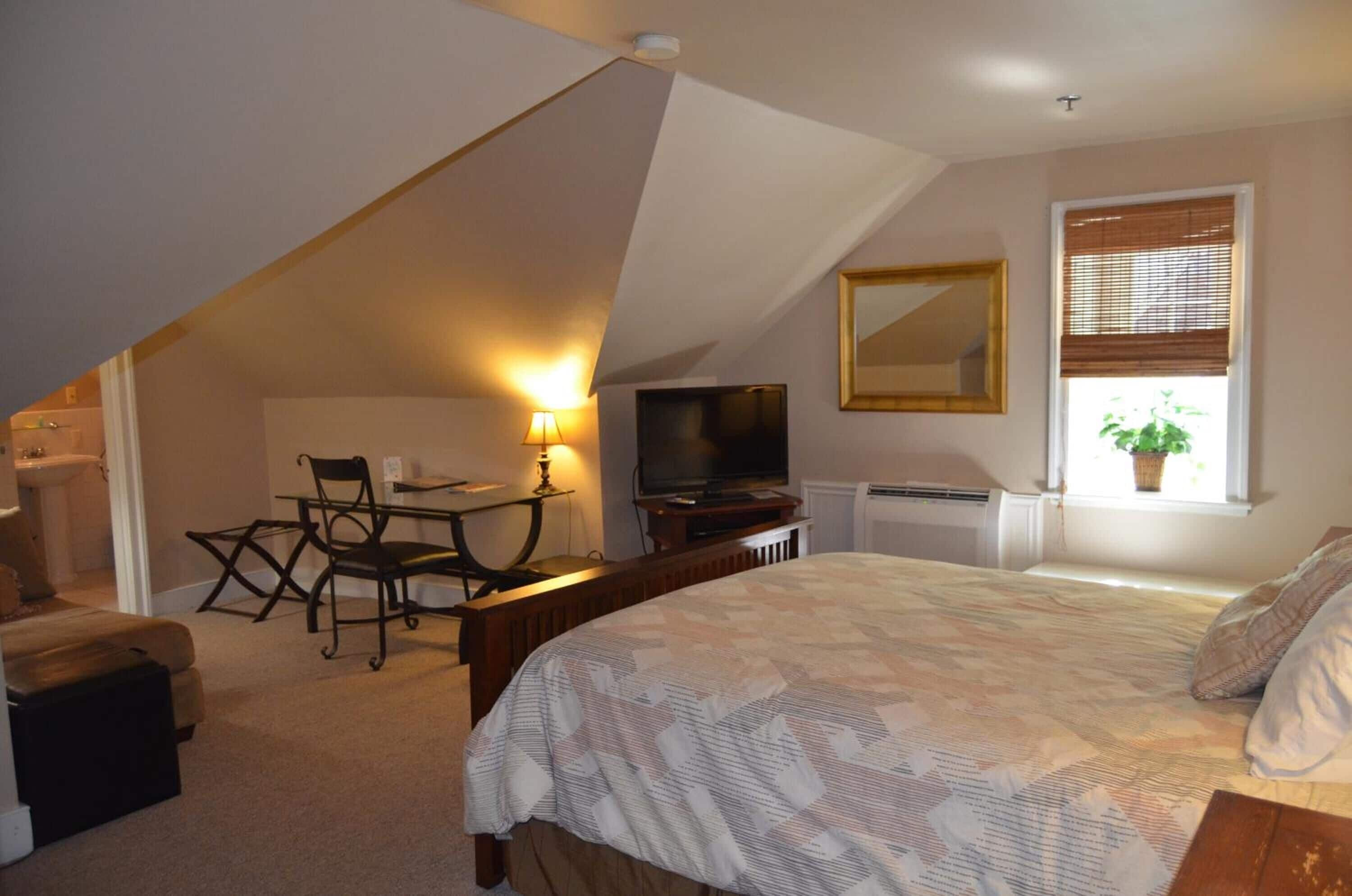 A bedroom with a bed, a desk with a chair, a television, and a window with blinds.