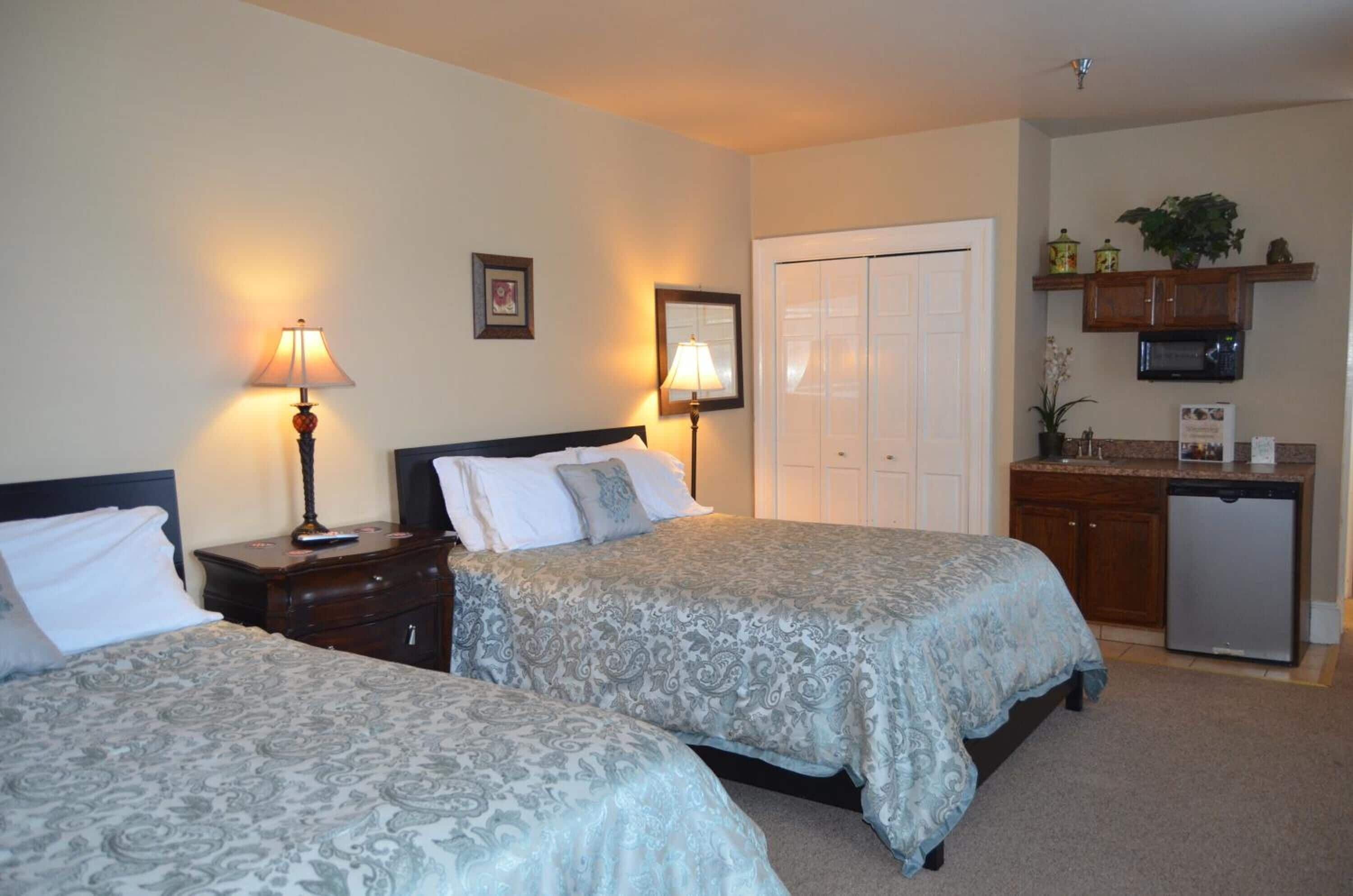 Une chambre d’hôtel avec deux lits, des lampes de chevet, une armoire, une petite kitchenette et une porte donnant sur une autre chambre.