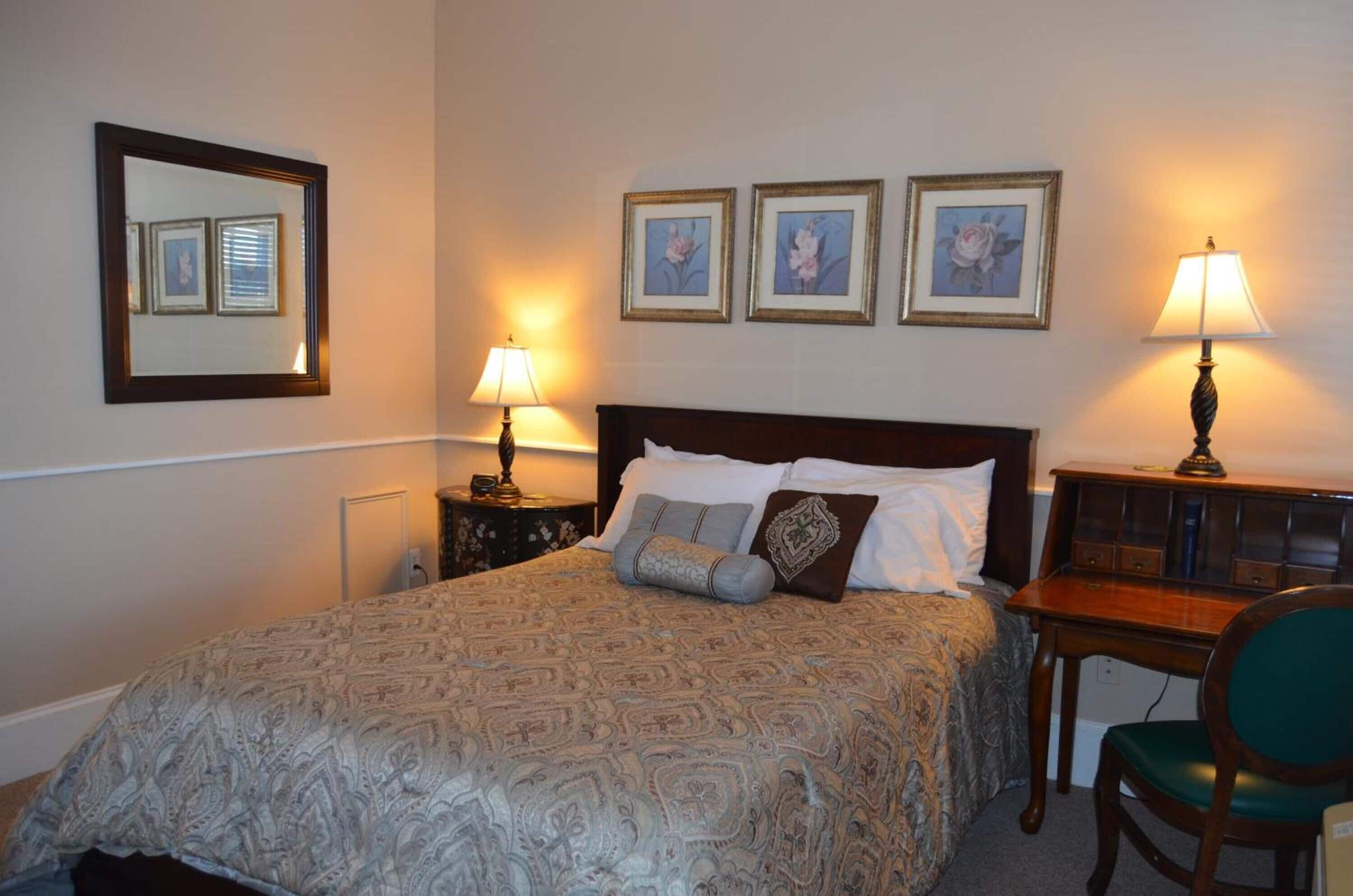 A neatly arranged hotel room with a large bed, two bedside tables with lamps, a desk with a chair, and three framed pictures on the wall.
