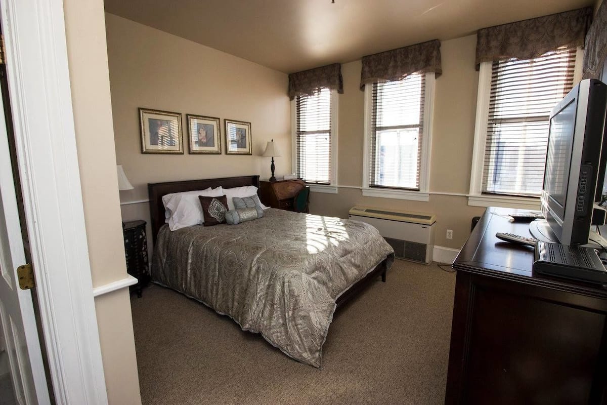 A bedroom with a bed, a desk with a computer and a TV, and three windows with curtains.