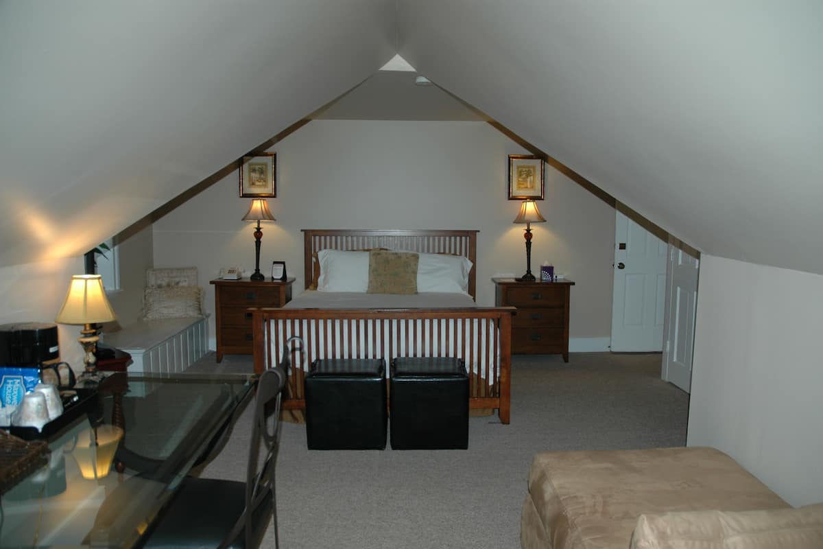 A bedroom with a sloped ceiling, a bed with a wooden headboard, two bedside tables with lamps, a bench, and a small table with a lamp.