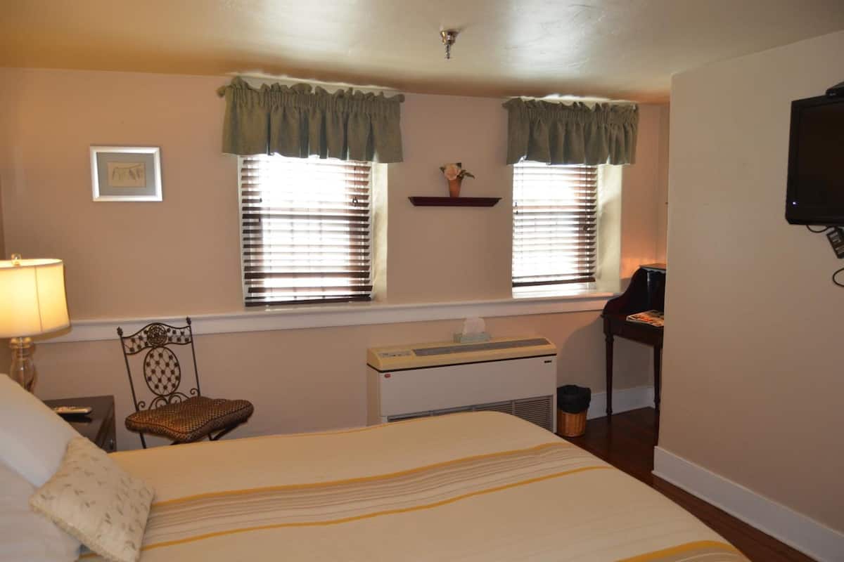 A hotel room with a bed, a chair, a desk with a computer, a heater, and a TV mounted on the wall.
