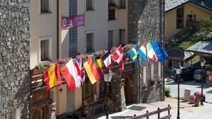 Un hotel de paredes de piedra, con varias ventanas y un letrero que dice "HOTEL". Hay banderas afuera y zonas de estar exteriores con mesas y sillas.
