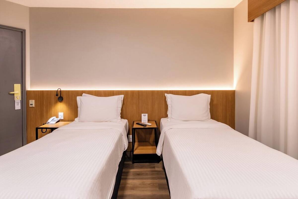 Una habitación de hotel con dos camas individuales, un cabecero de madera, una mesita de noche con lámpara y una puerta.