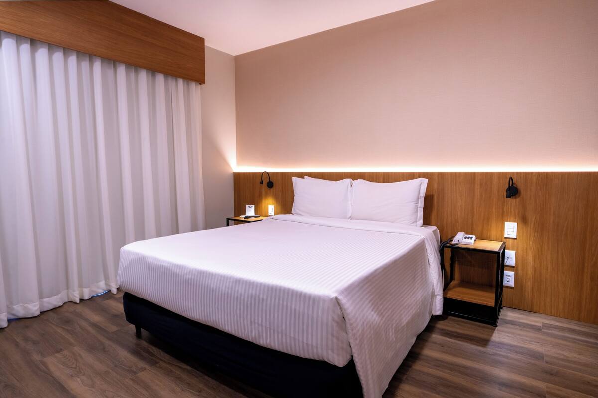 Una cama bien hecha con cabecera de madera, una mesita de noche con lámpara y un enchufe en la pared.
