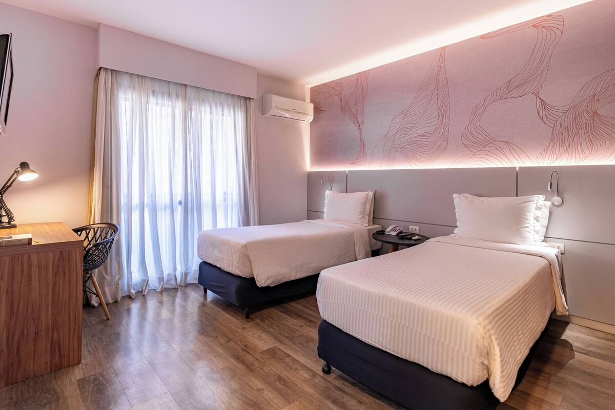 Habitación de hotel con dos camas, un escritorio, una silla y un ventanal con cortinas.
