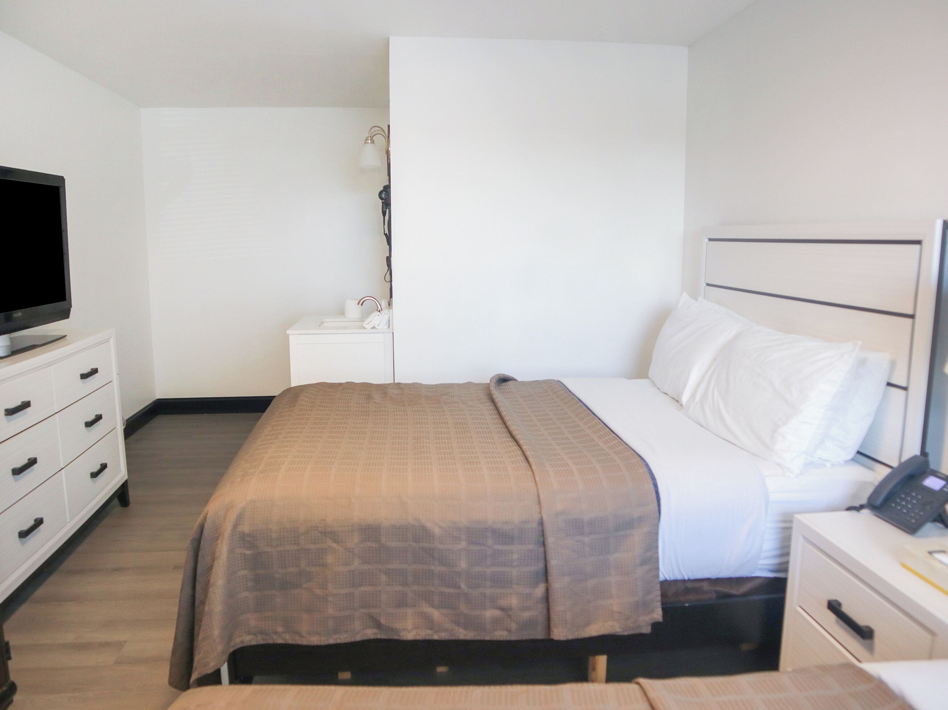 A hotel room with a bed, a TV on a dresser, a bedside table with a lamp, and a phone.